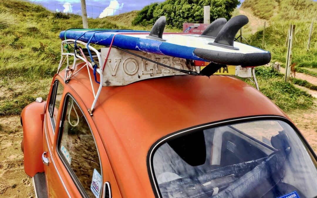 Oranje VW kever met een surfplank op zijn dak bij de duinen