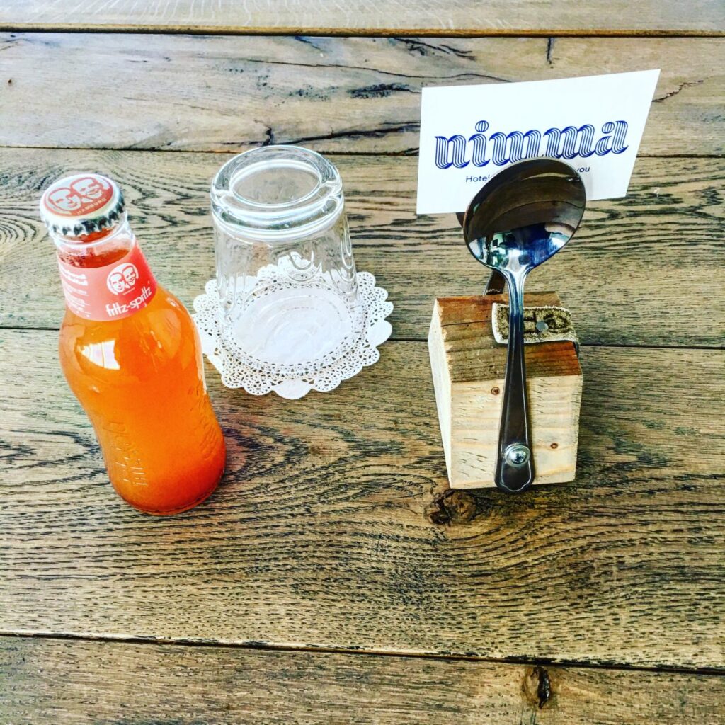 Houten tafel met flesje frisdrank erop en een rechtopstaande lepel met een kaartje erop met Nimma erop geschreven