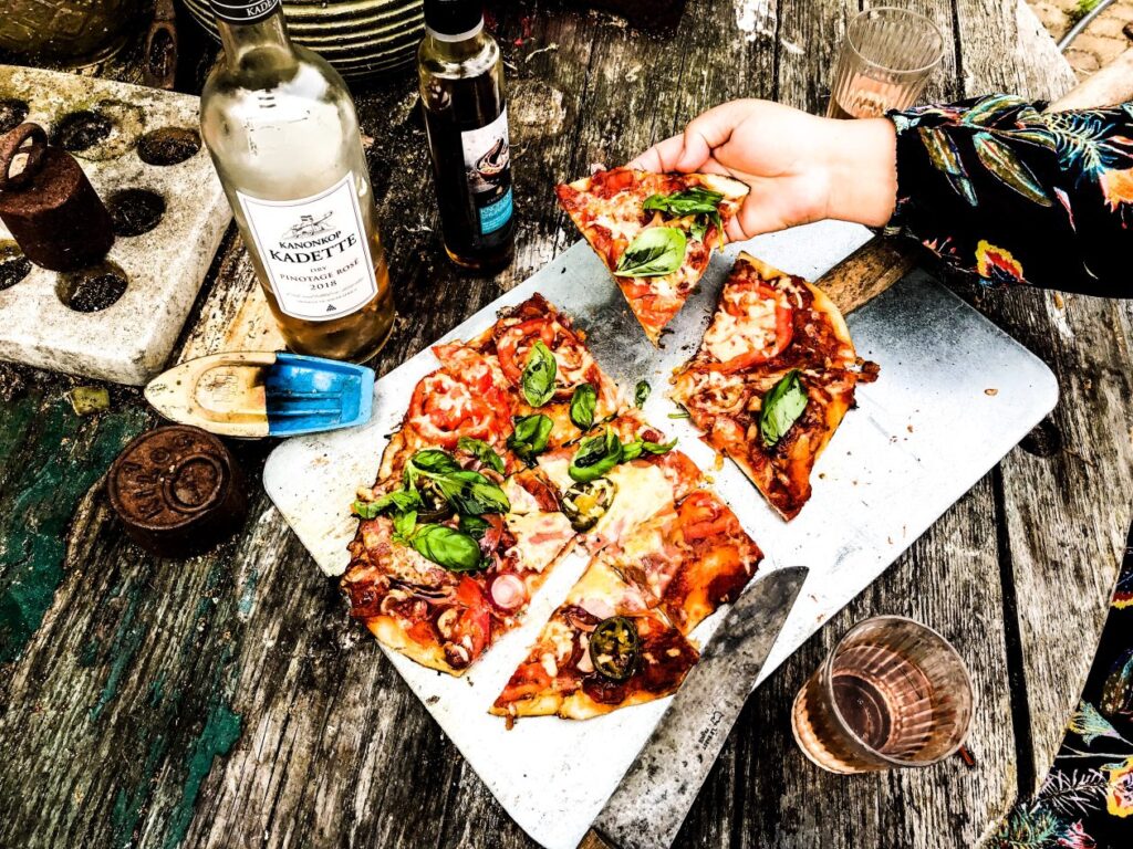houten tafel met daarop een snijplank met een pizza erop en een hand met een stuk pizza