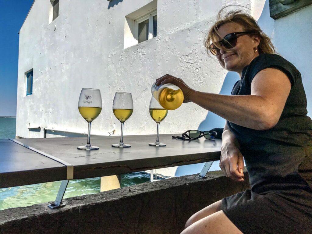 Vrouw schenk 4 glazen witte wijn in met uitzicht op een wit gebouw en de zee