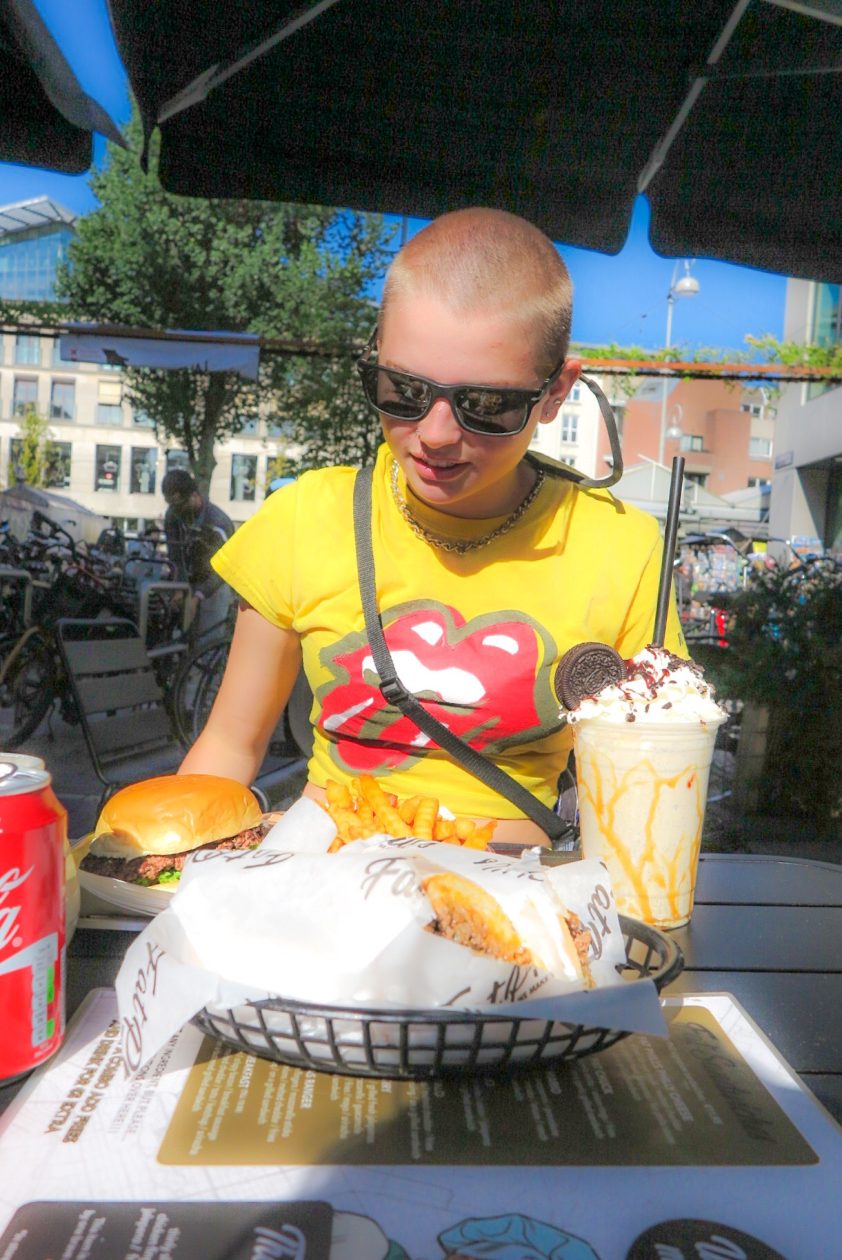 Meisje met kaal hoofd en geel t-Shirt aan , met voor haar een bord met fast food