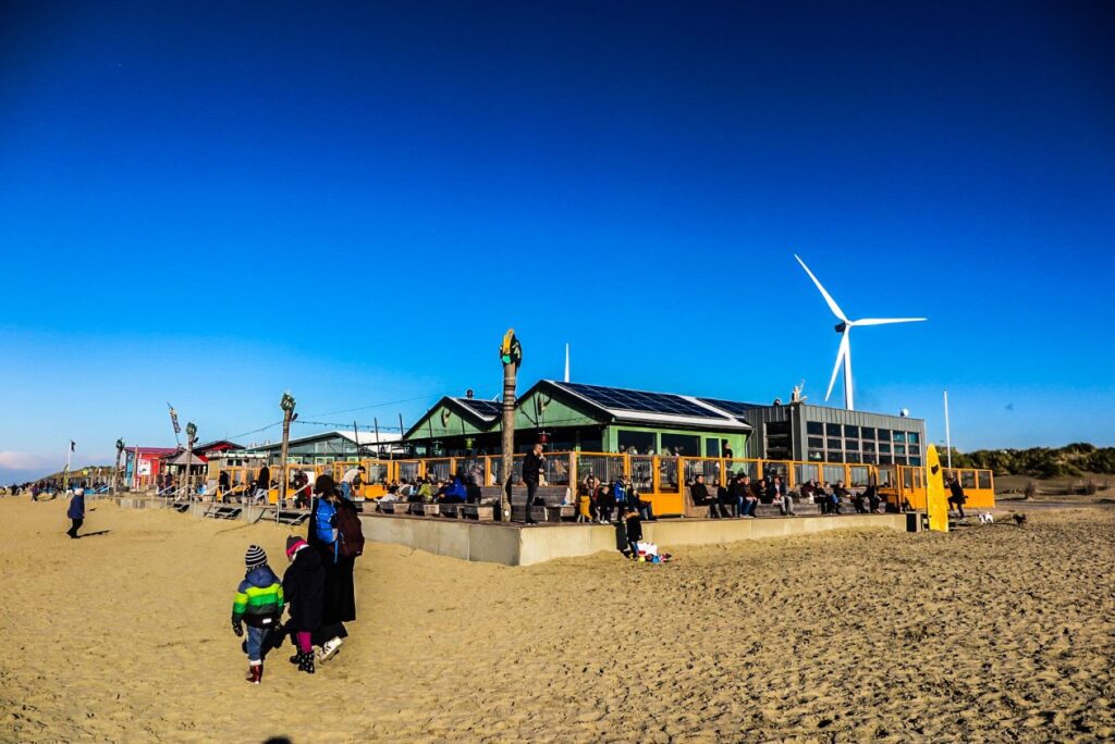 een strand paviljoen met daarachter een windmolen en op de voorgrond wandelende mensen