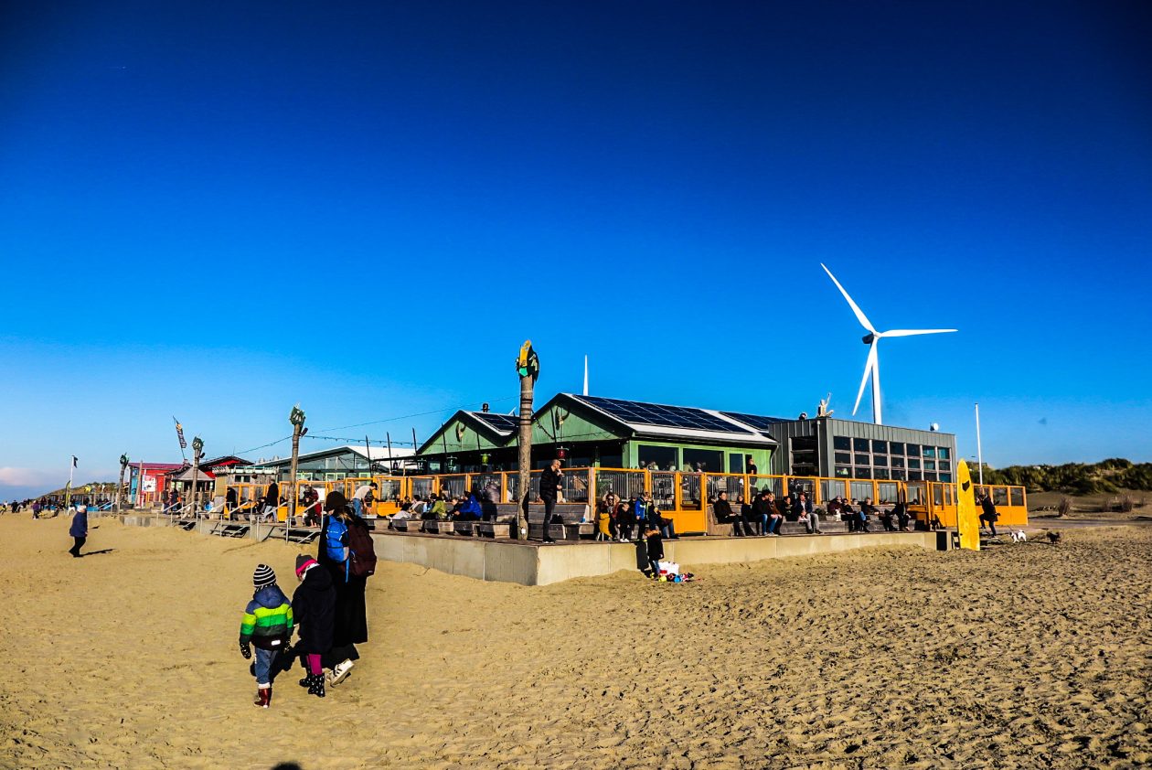 een strand paviljoen met daarachter een windmolen en op de voorgrond wandelende mensen