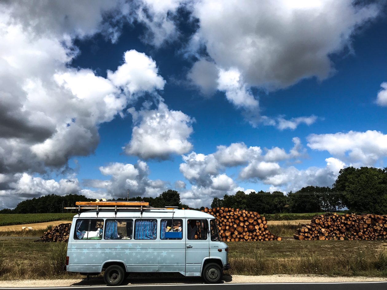 Blauwe camper met erachter opgestapelde houtblokken met een blauwe hemel met wolken 