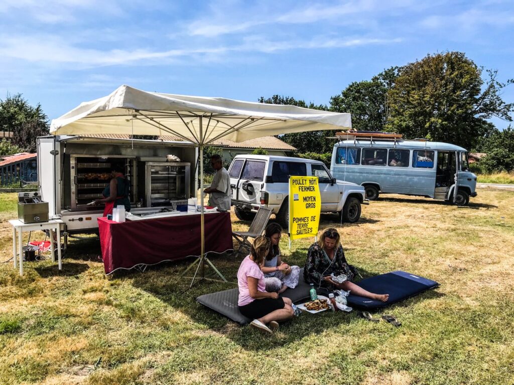 een foodtruck op het gras met vier mensen die op de grond zitten en in de verte een oude blauwe camper