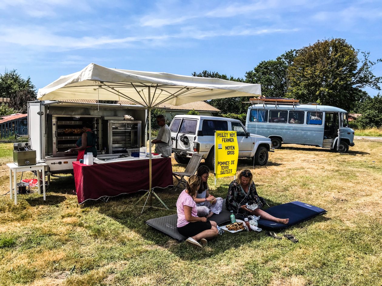 een foodtruck op het gras met vier mensen die op de grond zitten en in de verte een oude blauwe camper