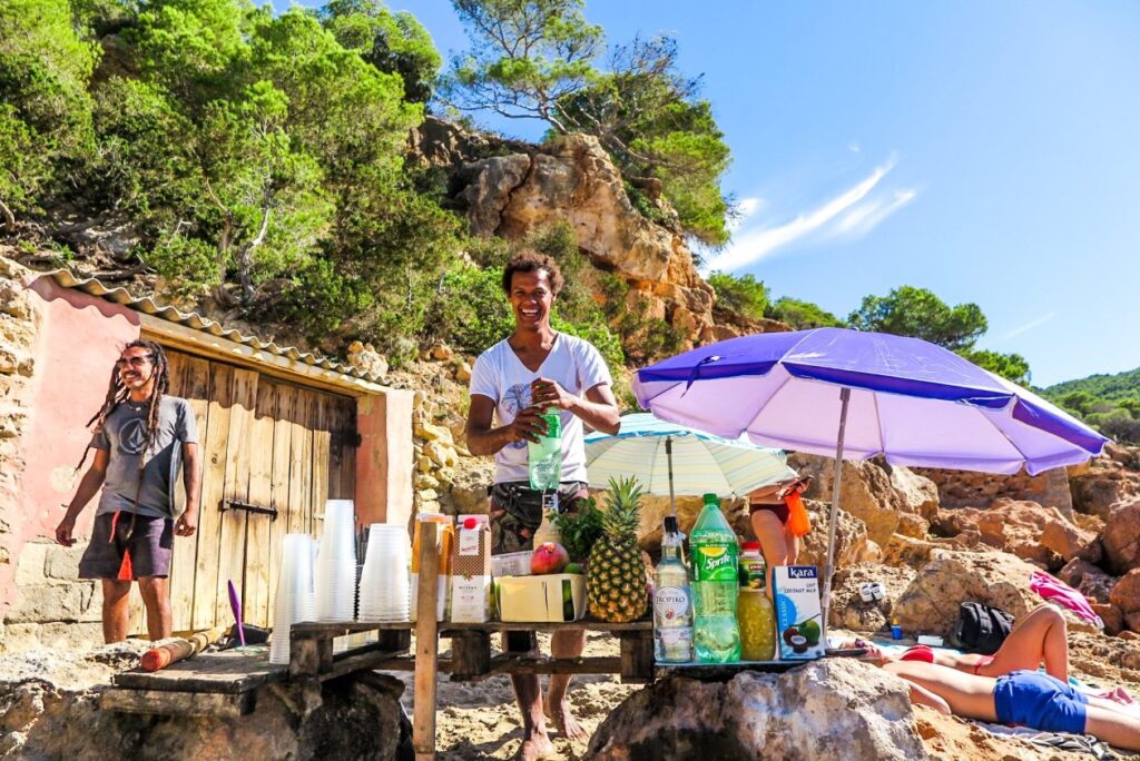 Lachende man staat recht op met een cocktail shaker op een strand met rechts een paarse parasol