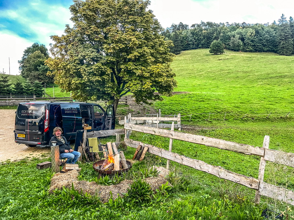 man bij een kampvuur , rechts loop een boeren hek met daarachter de weide